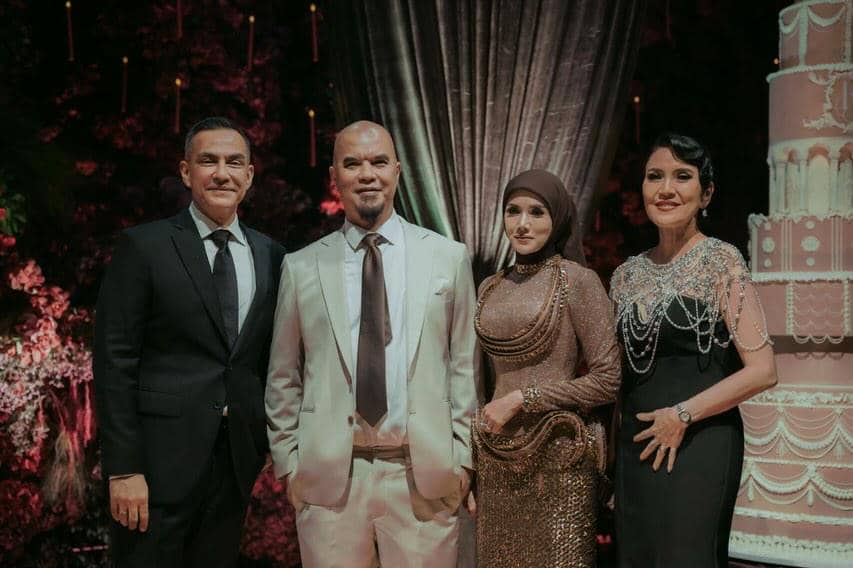  berpakaian formal berpose di acara pernikahan dengan latar dekorasi elegan dan kue pengantin besar di samping mereka.