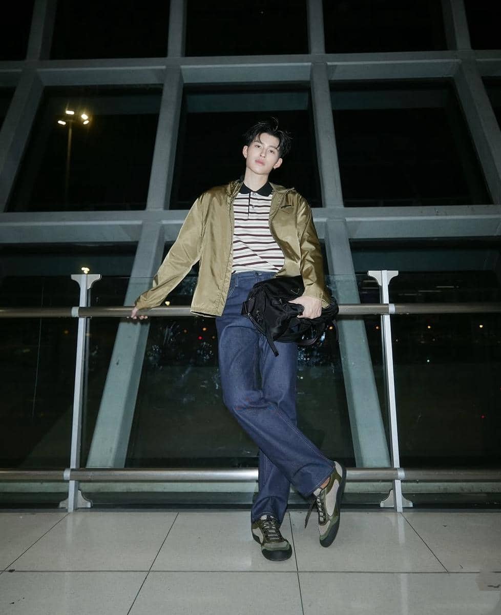 Seorang pria bergaya kasual mengenakan jaket emas, kaus bergaris, dan celana jeans berdiri di depan jendela besar pada malam hari.