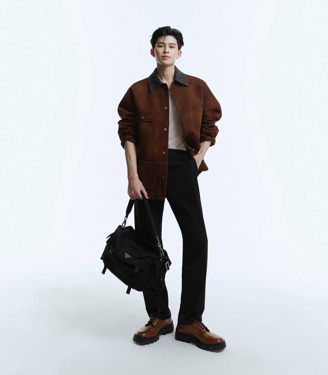 Seorang pria berpose dengan gaya modern mengenakan jaket cokelat, celana hitam, sepatu cokelat, dan membawa tas hitam di tangan.