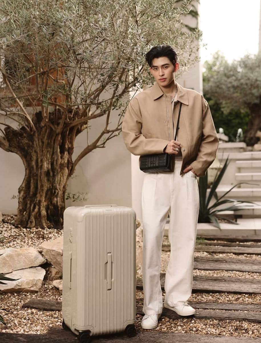 Pria berpenampilan stylish mengenakan jaket beige dan celana putih berdiri di luar ruangan dengan koper besar berwarna krem di sampingnya.