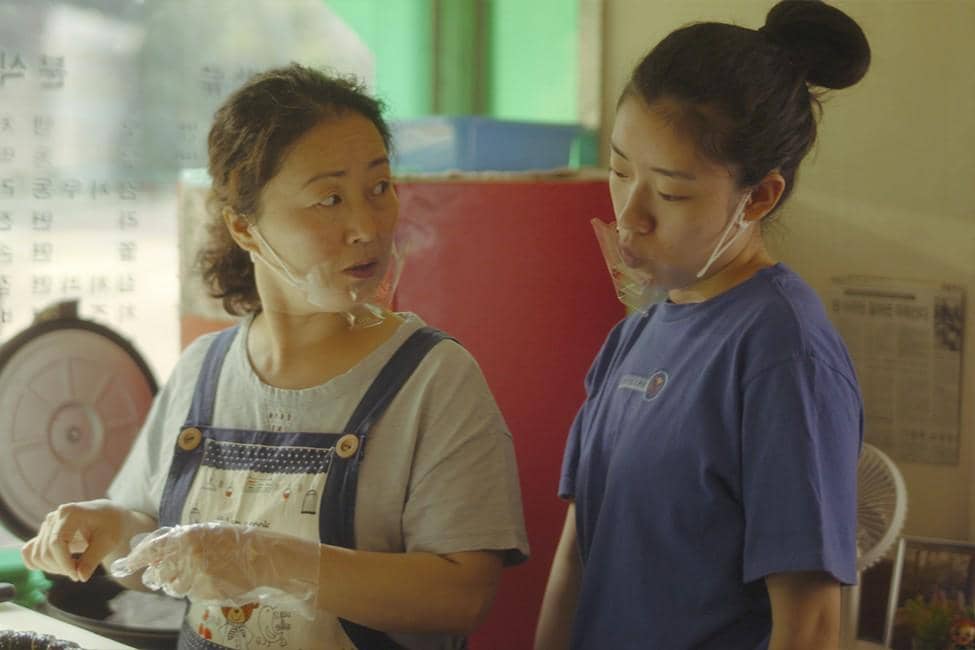 Dua wanita sedang berbicara di dapur kecil, salah satunya mengenakan celemek dan sarung tangan plastik sambil menyiapkan makanan.