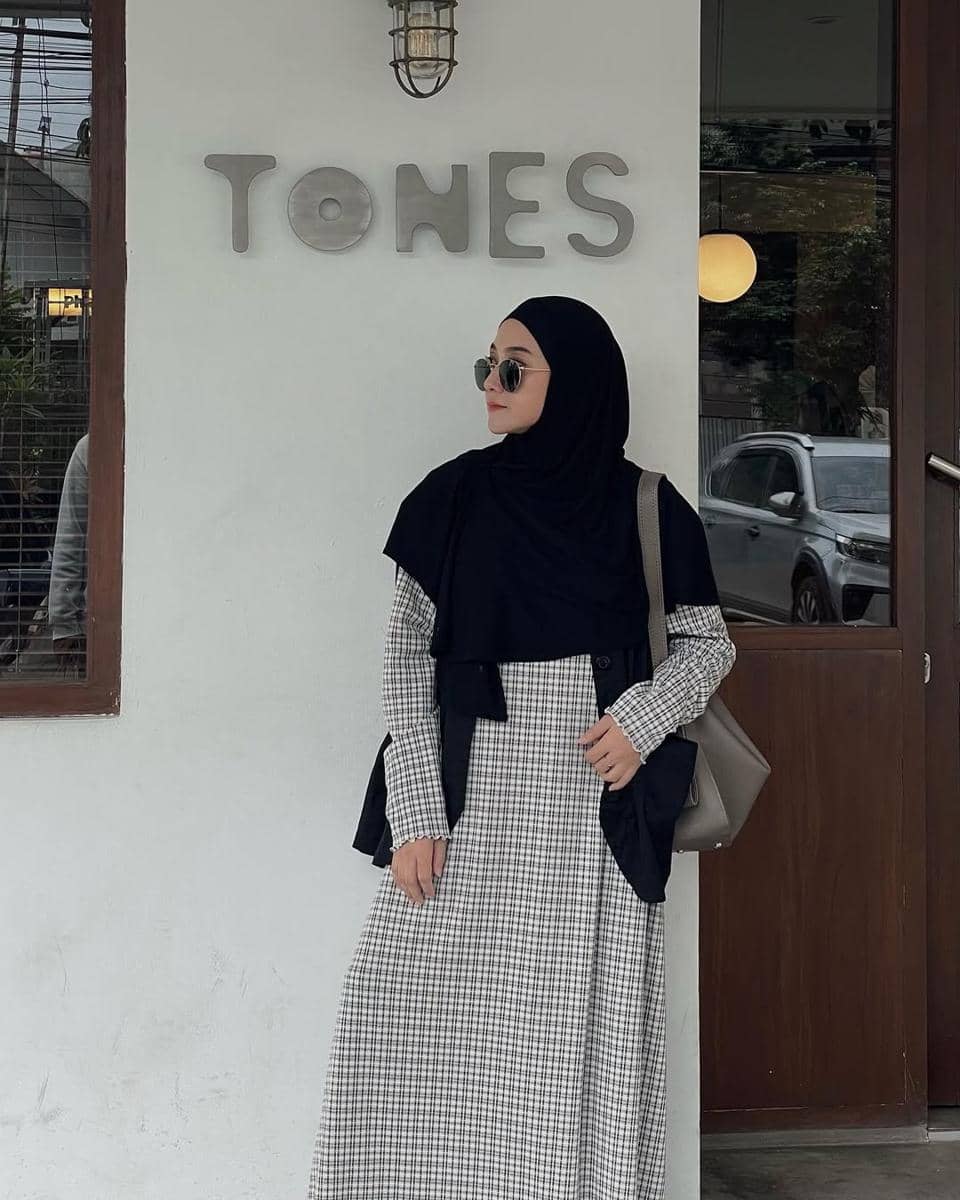 Seorang wanita berhijab hitam mengenakan gaun bermotif kotak-kotak berdiri di depan dinding bertuliskan TONES dengan gaya kasual.