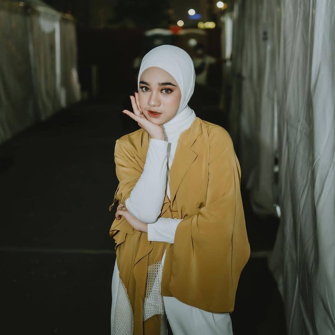 Seorang perempuan berhijab putih mengenakan outer mustard berpose dengan tangan di pipi di area luar ruangan pada malam hari.