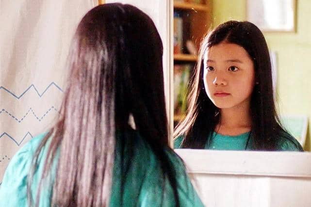 Seorang anak perempuan berambut panjang mengenakan kaus hijau sedang menatap pantulan dirinya di cermin dengan ekspresi serius.