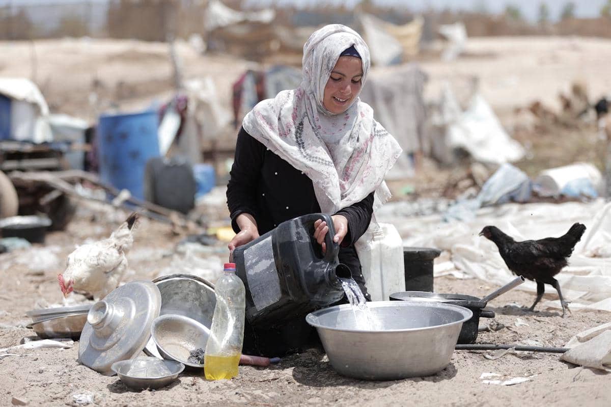 Perempuan berhijab di Gaza menuangkan air bersih ke dalam baskom logam di area terbuka dengan peralatan dapur di sekitarnya.