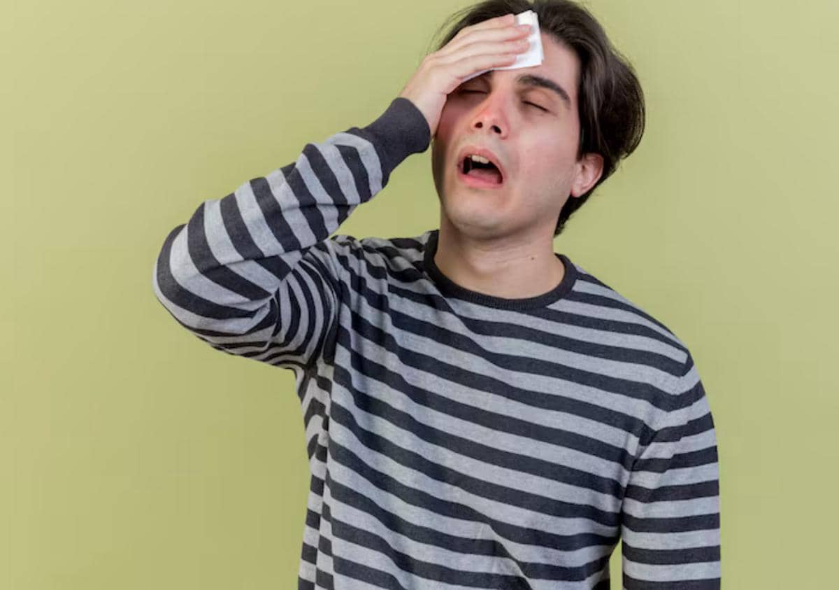 Seorang pria mengenakan kaus bergaris memegang dahinya dengan tisu, tampak lemah dan tidak enak badan di latar belakang hijau.