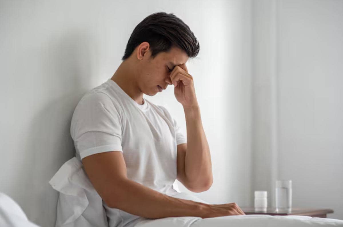 Seorang pria duduk di tempat tidur sambil memegang kepala, tampak lelah dan tidak enak badan di kamar bernuansa putih.