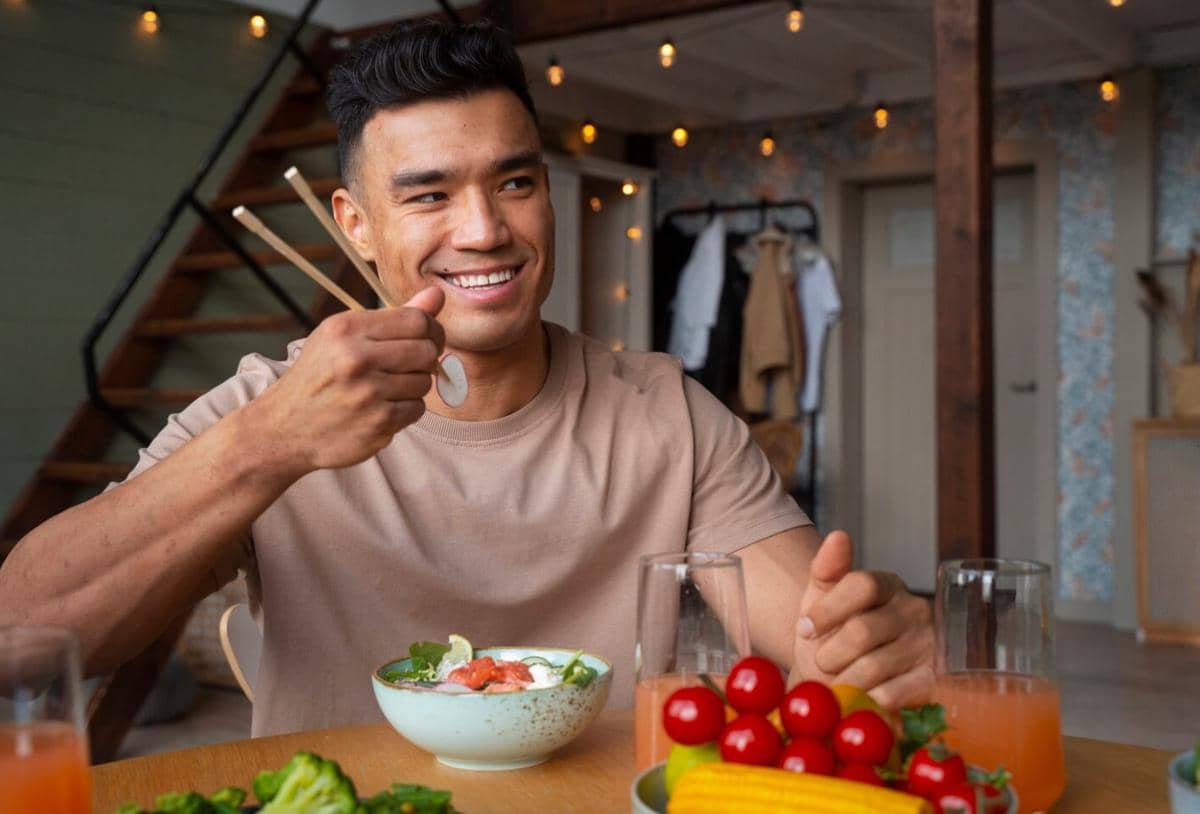 Seorang pria tersenyum sambil makan salad di meja kayu dengan sayuran segar dan jus di ruangan bernuansa hangat.