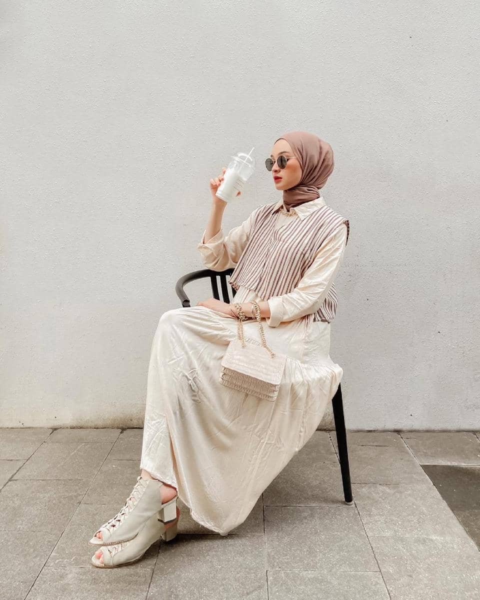 Seorang wanita berhijab duduk di kursi sambil memegang minuman, mengenakan busana berwarna krem dengan tas kecil dan kacamata hitam.