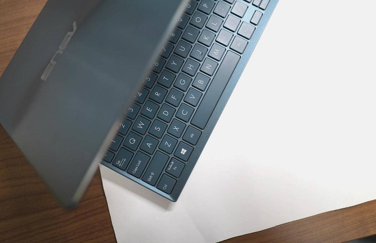 Keyboard laptop ASUS berwarna abu-abu tua terlihat sebagian di atas meja kayu dengan latar kertas putih di bawahnya.
