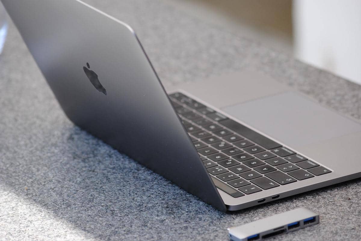 Laptop MacBook Pro berwarna abu-abu terbuka di atas meja granit dengan adaptor USB di sampingnya.
