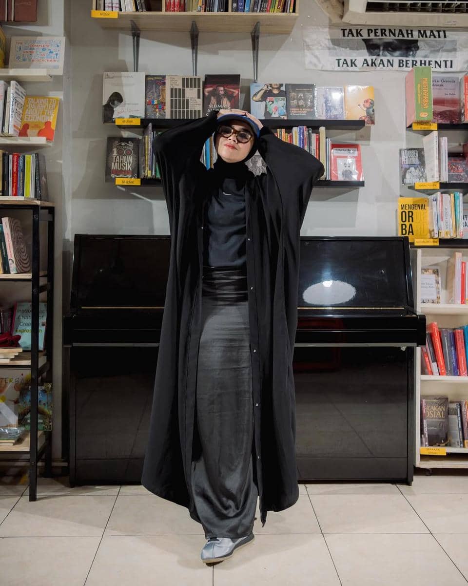 Seorang perempuan mengenakan busana serba hitam dengan kacamata hitam berdiri di depan rak buku dan piano di sebuah toko buku.
