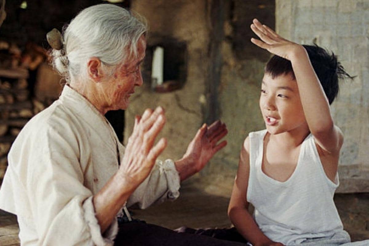 Seorang nenek berambut putih tersenyum sambil bermain tangan dengan seorang anak laki-laki kecil di dalam rumah sederhana.