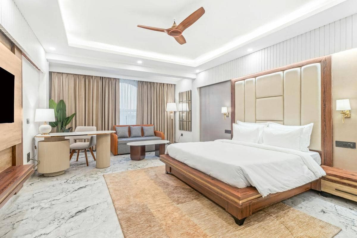Kamar tidur modern dengan tempat tidur kayu besar, pencahayaan hangat, meja kerja, dan sofa kecil di dekat jendela ber tirai panjang.