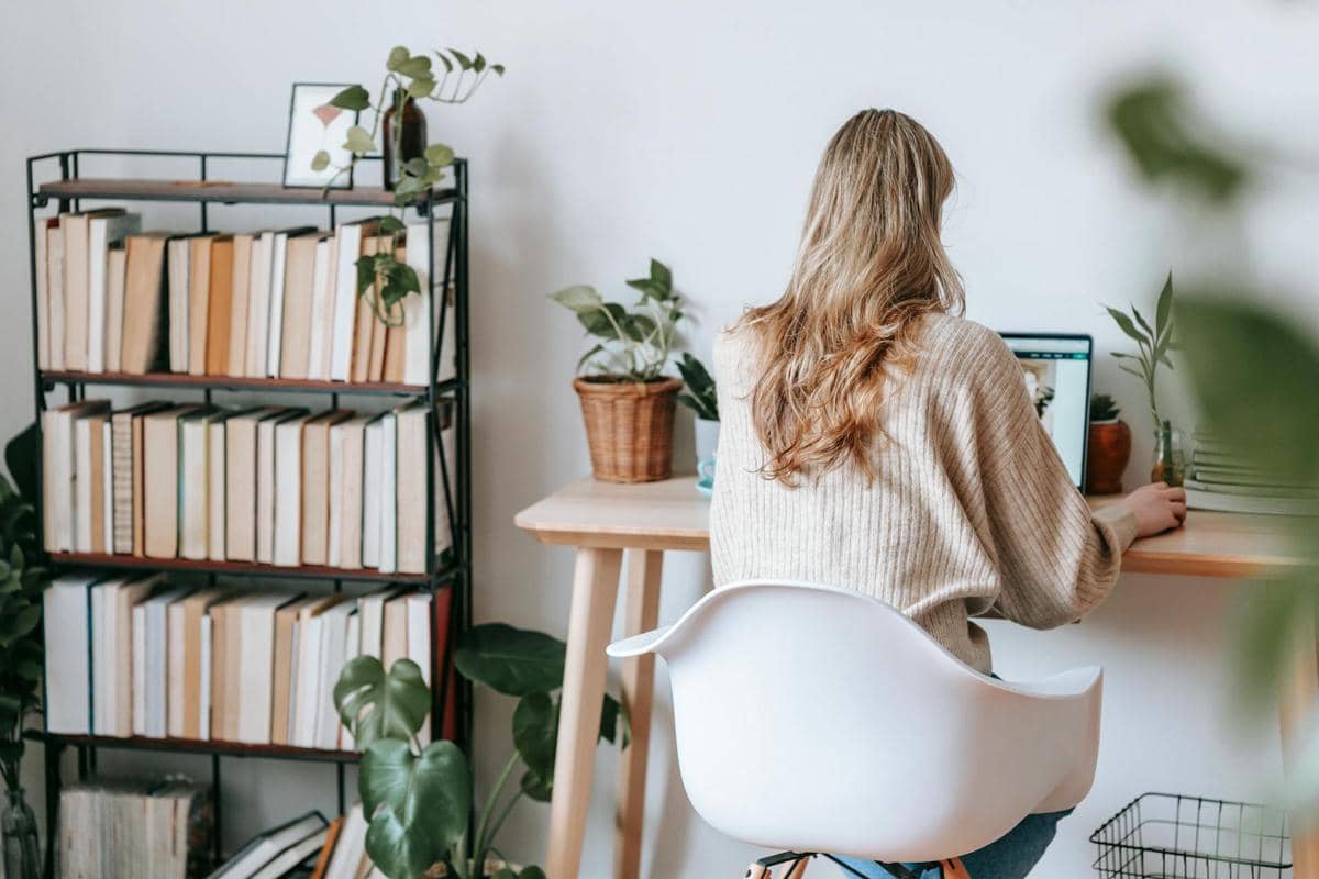 Seorang perempuan duduk di meja kerja dengan laptop di ruang baca minimalis yang dilengkapi rak buku dan tanaman hias.
