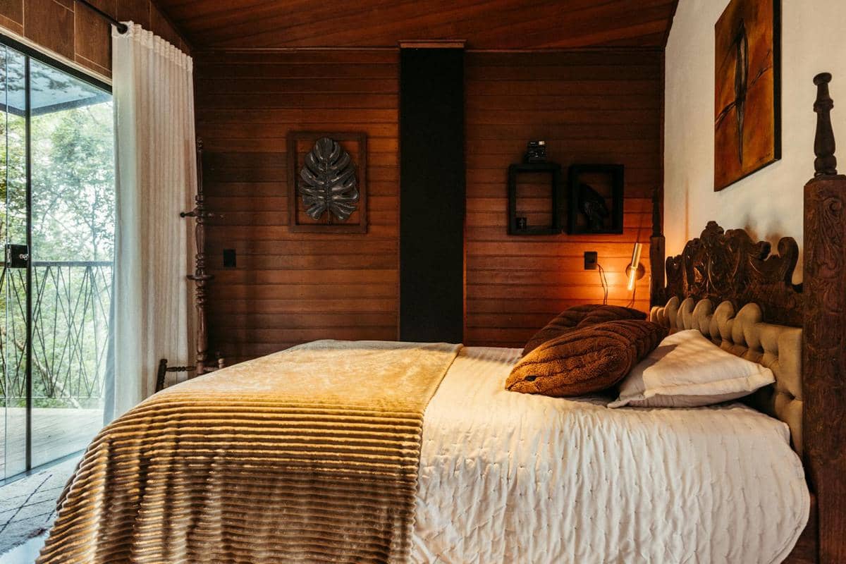 Kamar tidur bergaya rustic dengan dinding kayu, tempat tidur berukir, selimut cokelat, dan pencahayaan hangat di dekat jendela kaca besar.