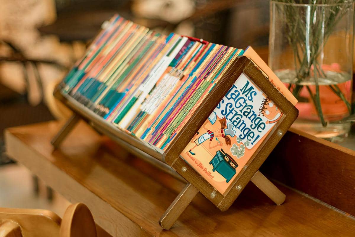 Rak buku minimalis dari kayu berisi deretan buku warna-warni tersusun rapi di atas meja kayu di ruang baca yang hangat.