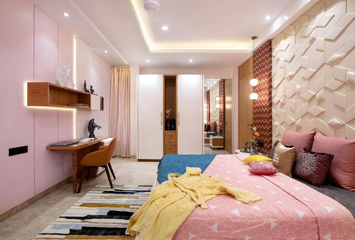 Kamar tidur modern dengan dinding bertekstur, tempat tidur berseprai pink, meja kerja kayu, dan pencahayaan hangat yang elegan.