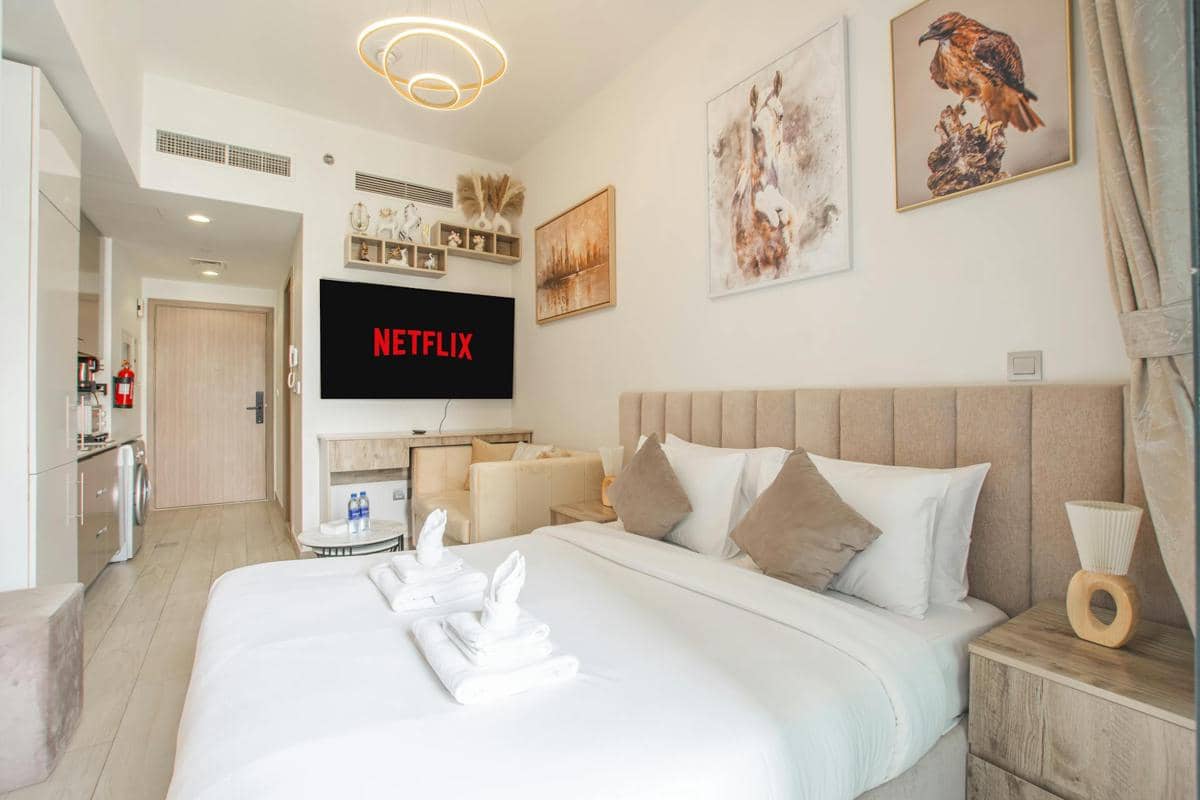 Kamar tidur modern dengan tempat tidur rapi, dekorasi netral, televisi besar menampilkan logo Netflix, dan pencahayaan elegan.