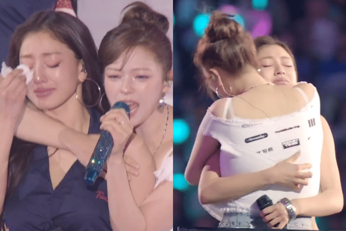 Dua anggota grup musik perempuan sedang berpelukan di atas panggung konser, salah satunya tampak menangis sambil memegang mikrofon.