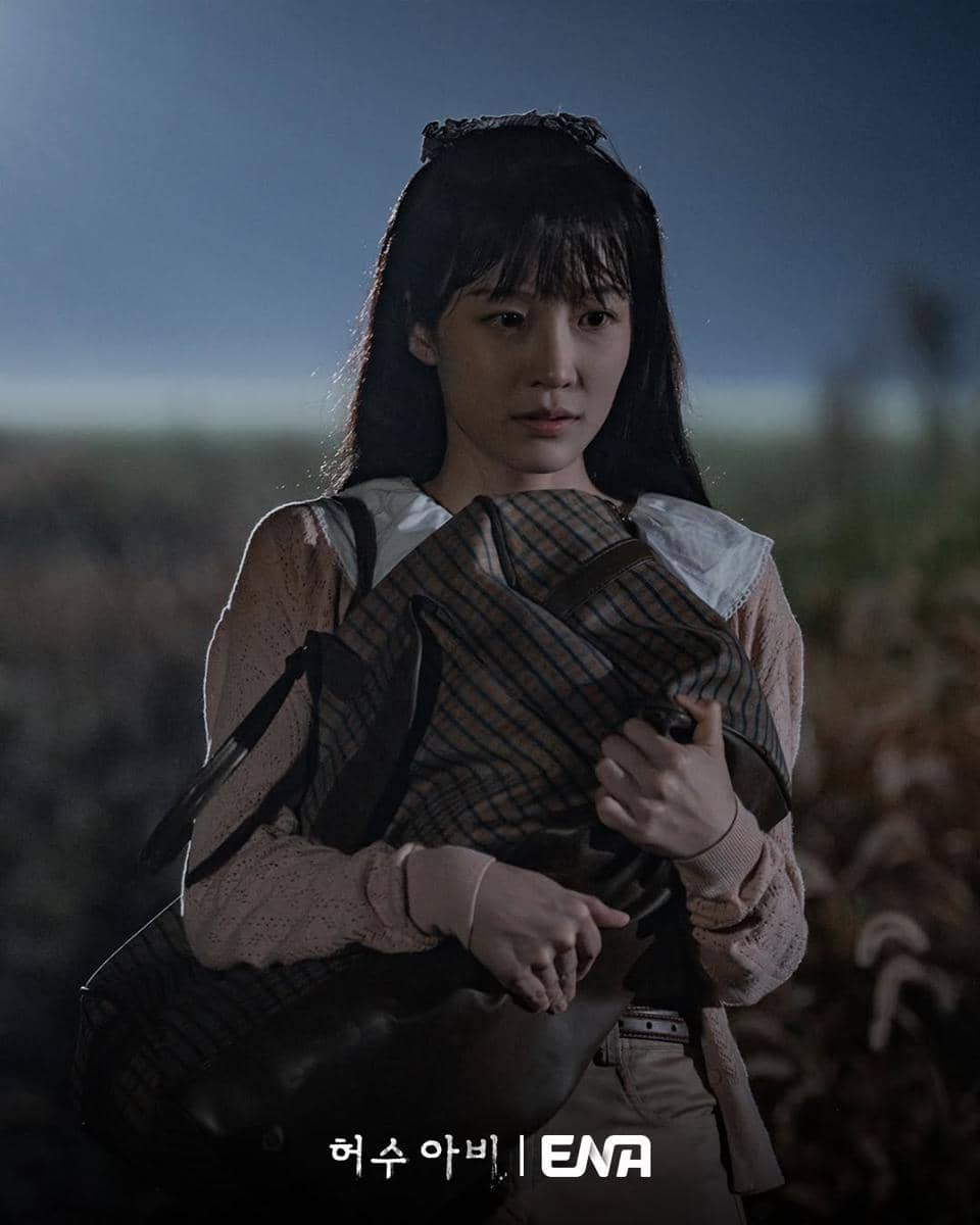 Seorang perempuan muda berdiri di luar ruangan pada malam hari sambil memeluk tas besar, dengan latar ladang gelap di belakangnya.