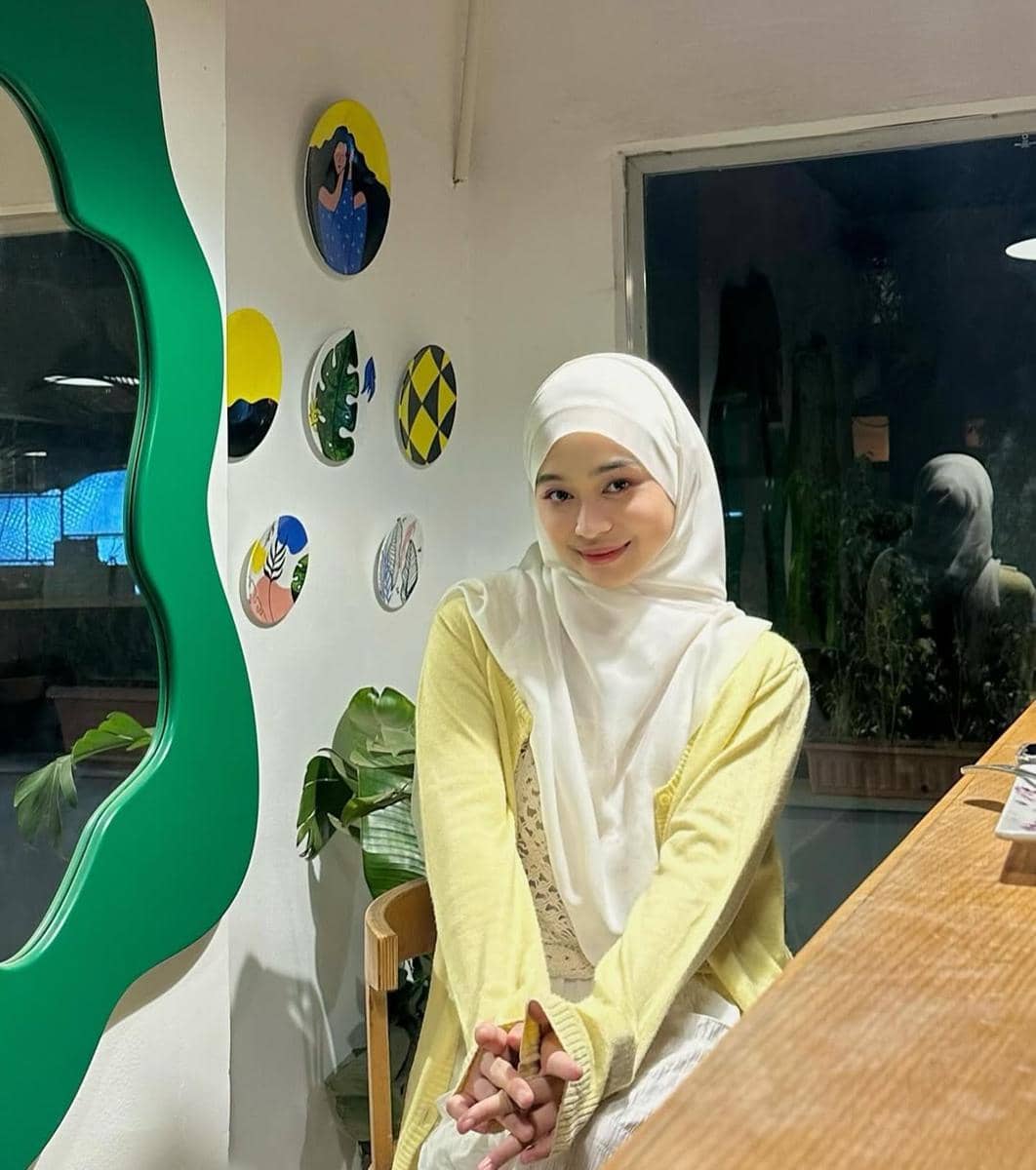 Seorang perempuan berhijab putih dan mengenakan cardigan kuning duduk di dekat meja kayu dengan latar dinding berdekorasi seni.