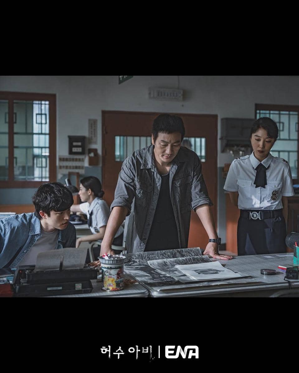 Tiga orang berdiskusi serius di ruang kantor dengan dokumen dan foto di atas meja, suasana tegang menggambarkan adegan investigasi.