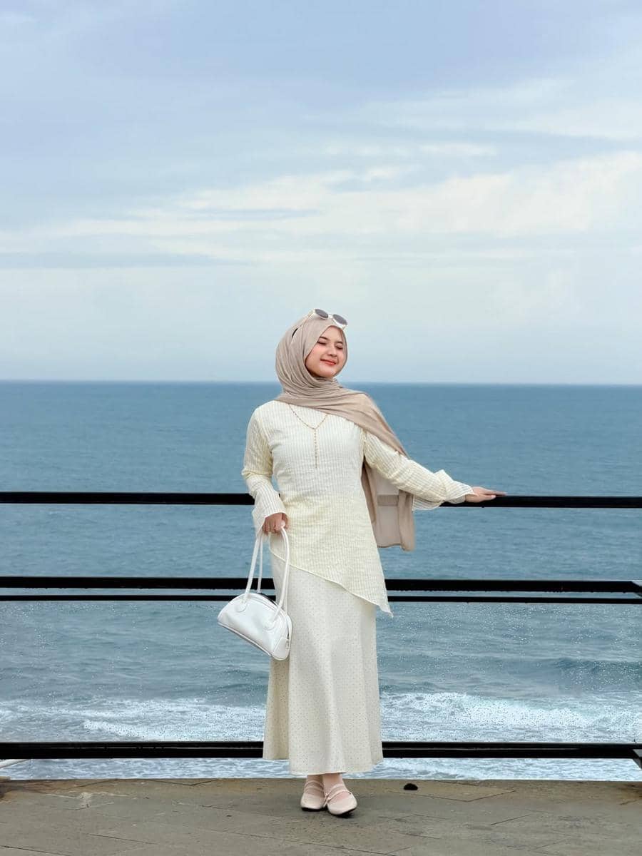 Seorang wanita berhijab berpose di tepi laut dengan latar belakang ombak dan langit cerah, mengenakan busana berwarna krem dan membawa tas putih.