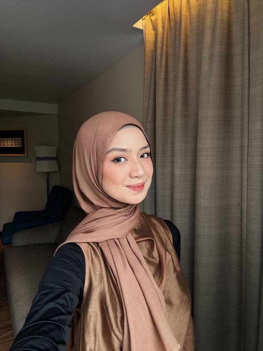 Seorang perempuan berhijab cokelat tersenyum di dalam ruangan dengan tirai dan sofa di latar belakang.