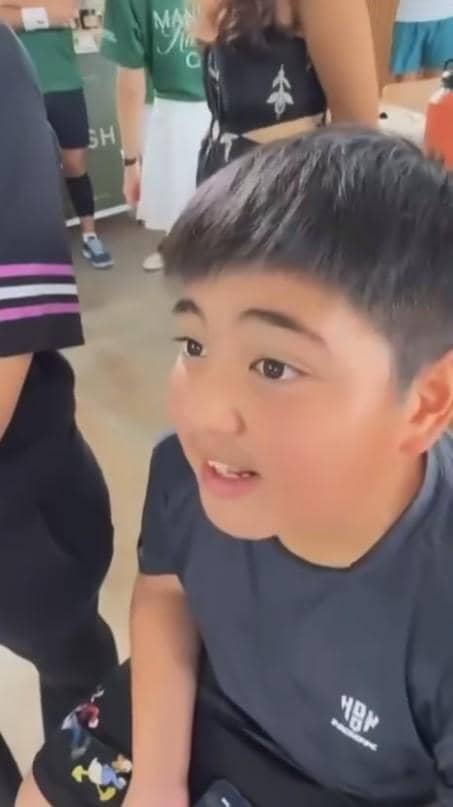 Seorang anak laki-laki mengenakan kaus hitam sedang duduk di area turnamen padel di Singapura bersama beberapa orang di sekitarnya.