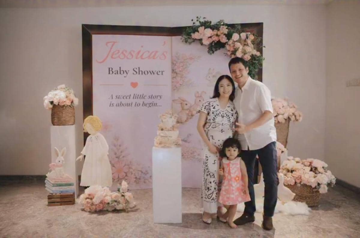Sebuah keluarga berpose di acara baby shower dengan dekorasi bunga berwarna pastel dan latar bertuliskan 'Jessica’s Baby Shower'.