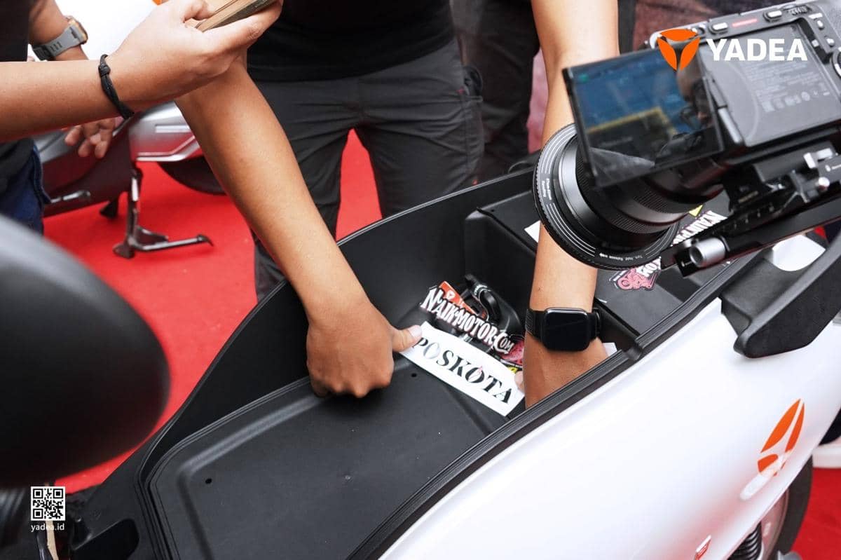 Seseorang memeriksa dan menyegel baterai di bagasi motor listrik Yadea dengan latar belakang karpet merah dan kamera merekam prosesnya.