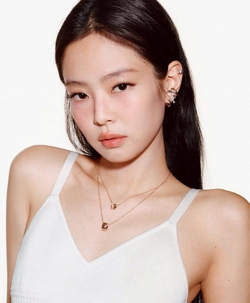 Seorang perempuan berambut panjang mengenakan atasan putih tanpa lengan dan kalung emas ganda, berpose di latar belakang putih.