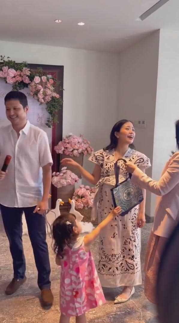 Seorang wanita hamil tersenyum bersama seorang pria dan anak kecil di acara baby shower dengan dekorasi bunga berwarna merah muda.