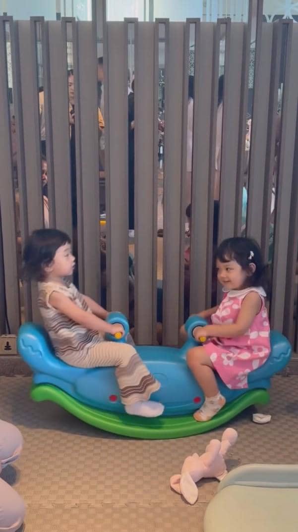 Dua anak kecil bermain di mainan jungkat-jungkit biru saat acara baby shower di dalam ruangan dengan suasana hangat dan ramai.