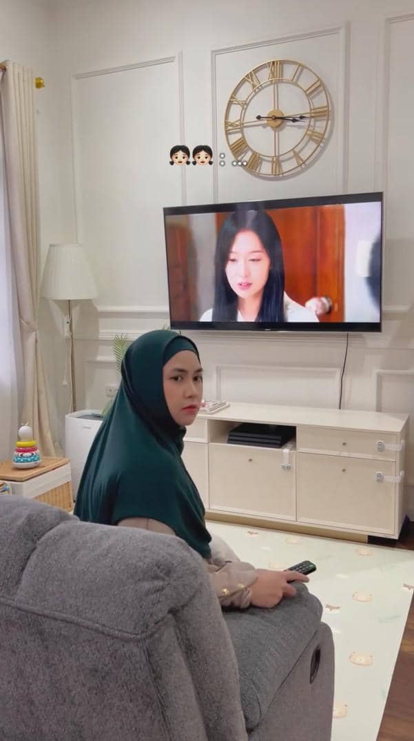 Seorang wanita berhijab duduk di sofa ruang tamu modern sambil memegang remote dan menonton televisi di dinding.