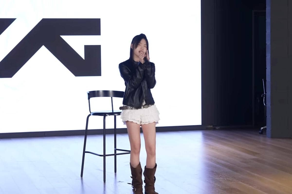 Seorang peserta pelatihan berdiri di atas panggung kayu dengan latar belakang logo besar berwarna hitam dan putih, mengenakan jaket kulit hitam dan rok putih.