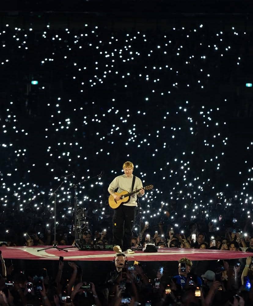 Seorang musisi memainkan gitar di atas panggung besar dengan lautan penonton menyalakan lampu ponsel di sekitarnya.