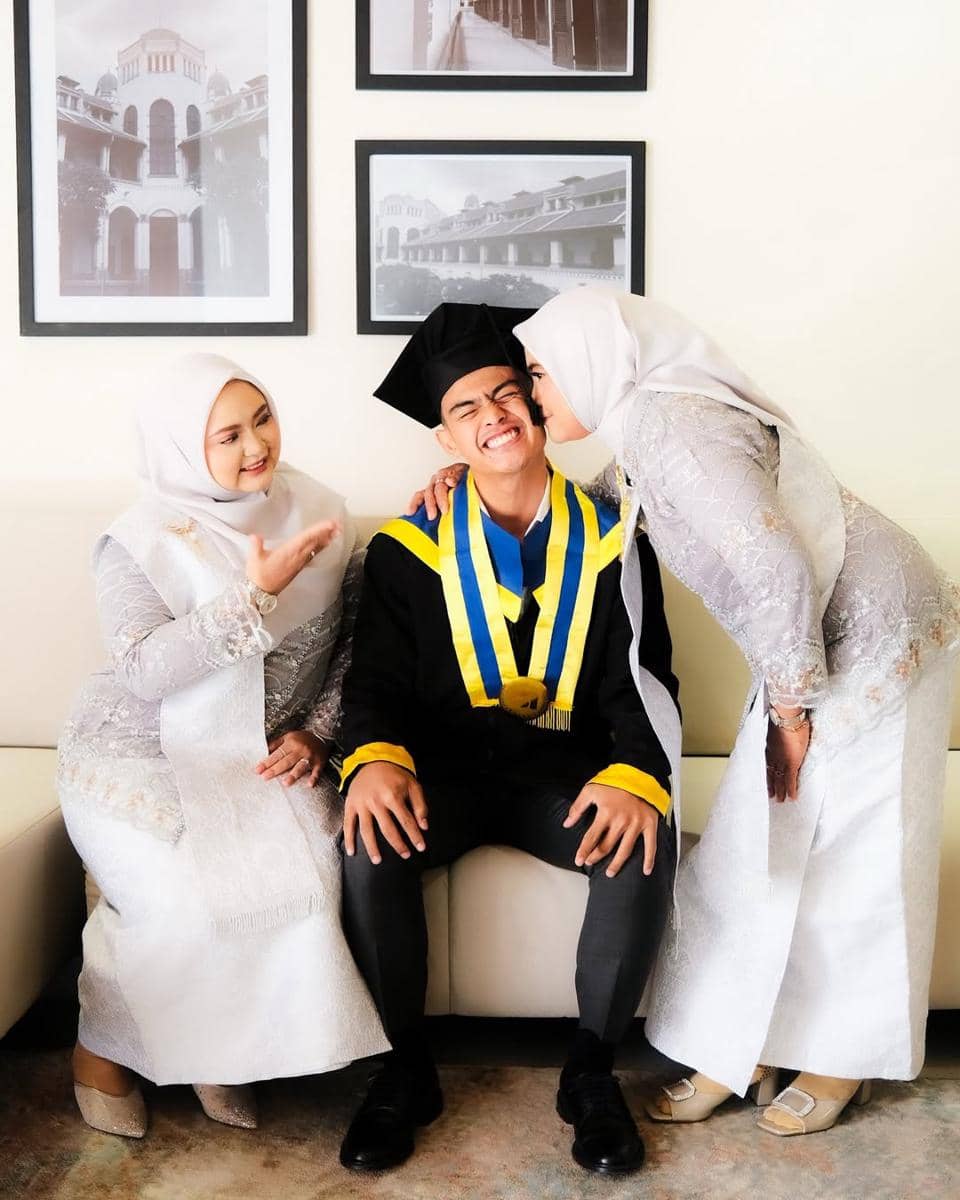 Pratama Arhan mengenakan toga hitam dan selempang kuning biru tersenyum saat dirayakan bersama dua wanita berhijab berpakaian kebaya putih.