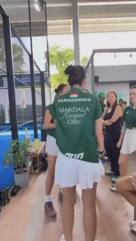 Seorang perempuan mengenakan seragam hijau bertuliskan Mandala Racquet Club berbincang dengan peserta lain di area lapangan padel dalam ruangan.