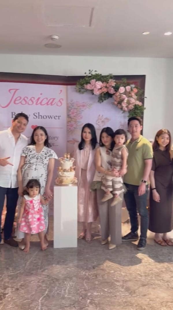 Beberapa orang berpose bersama di acara baby shower dengan dekorasi bunga dan kue bertingkat di latar belakang bertuliskan Jessica's Baby Shower.