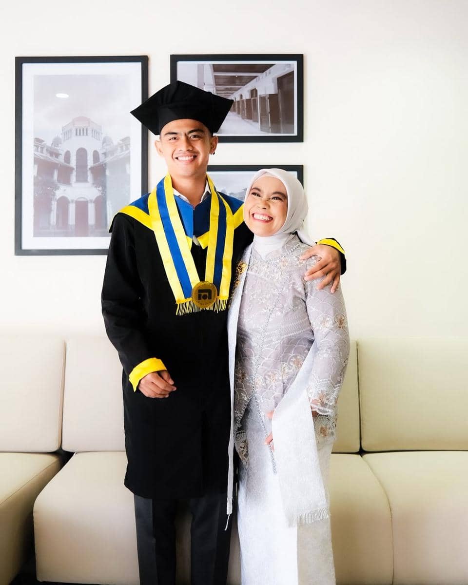 Seorang wisudawan mengenakan toga hitam dengan selempang biru dan kuning berpose bersama seorang wanita berhijab di ruang tamu.