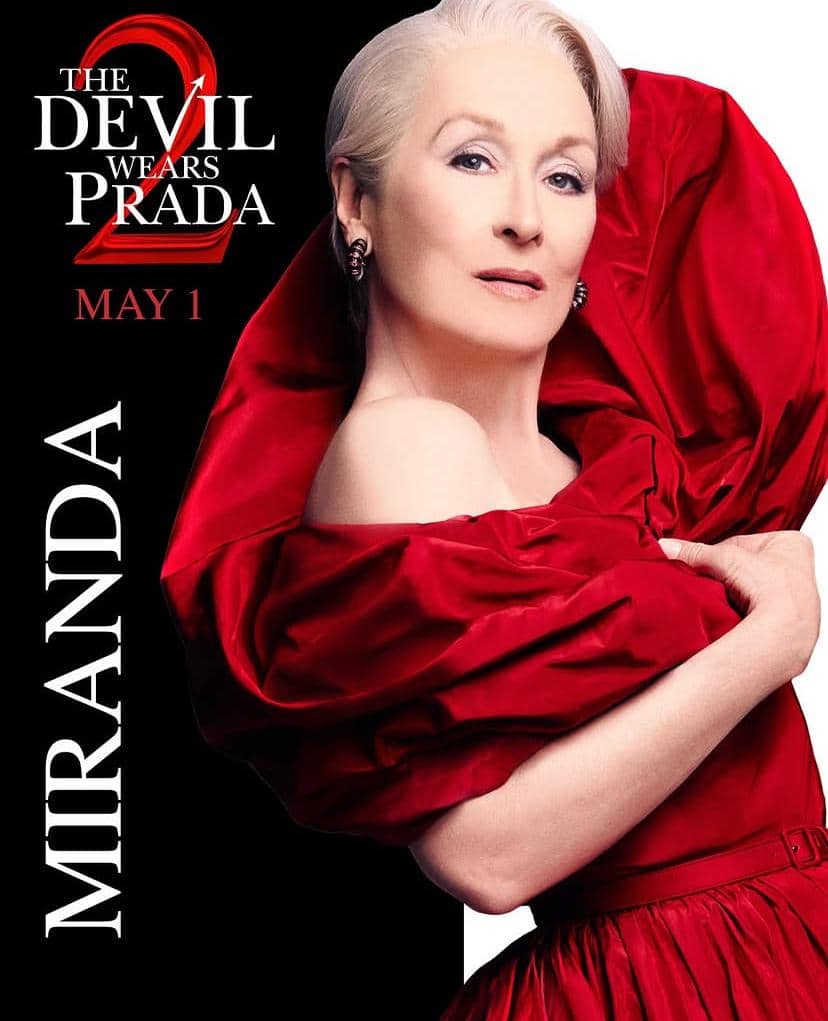 Seorang perempuan berambut putih mengenakan gaun merah elegan dengan latar promosi film The Devil Wears Prada 2 yang menampilkan tulisan tanggal rilis.