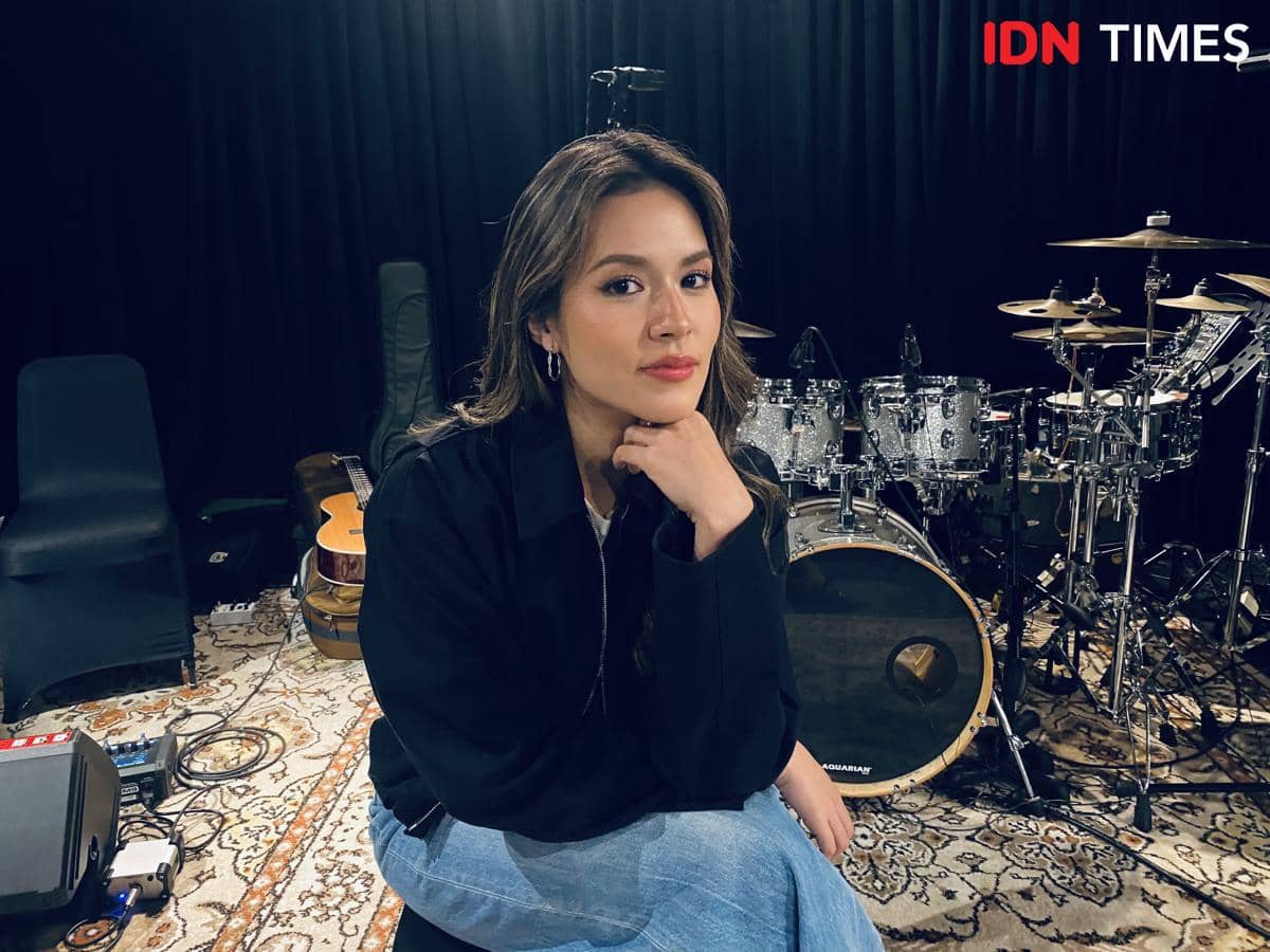 Seorang perempuan duduk di studio musik dengan latar drum, gitar, dan peralatan musik lainnya, mengenakan jaket hitam dan celana jeans.