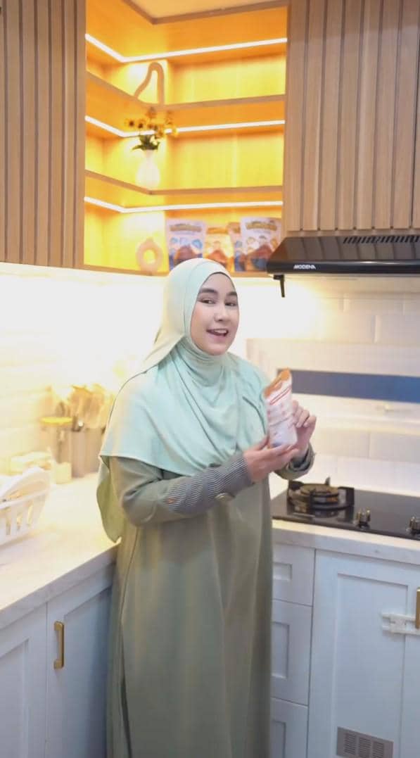 Seorang wanita berhijab berdiri di dapur modern berwarna putih dan kayu, memegang kemasan produk di dekat kompor gas.