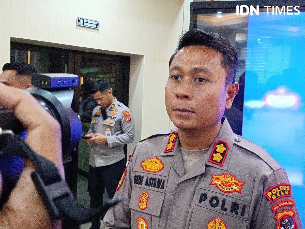 Seorang perwira polisi berpakaian dinas lengkap berdiri di dalam ruangan, dengan latar belakang layar biru dan beberapa petugas lain di sekitarnya.
