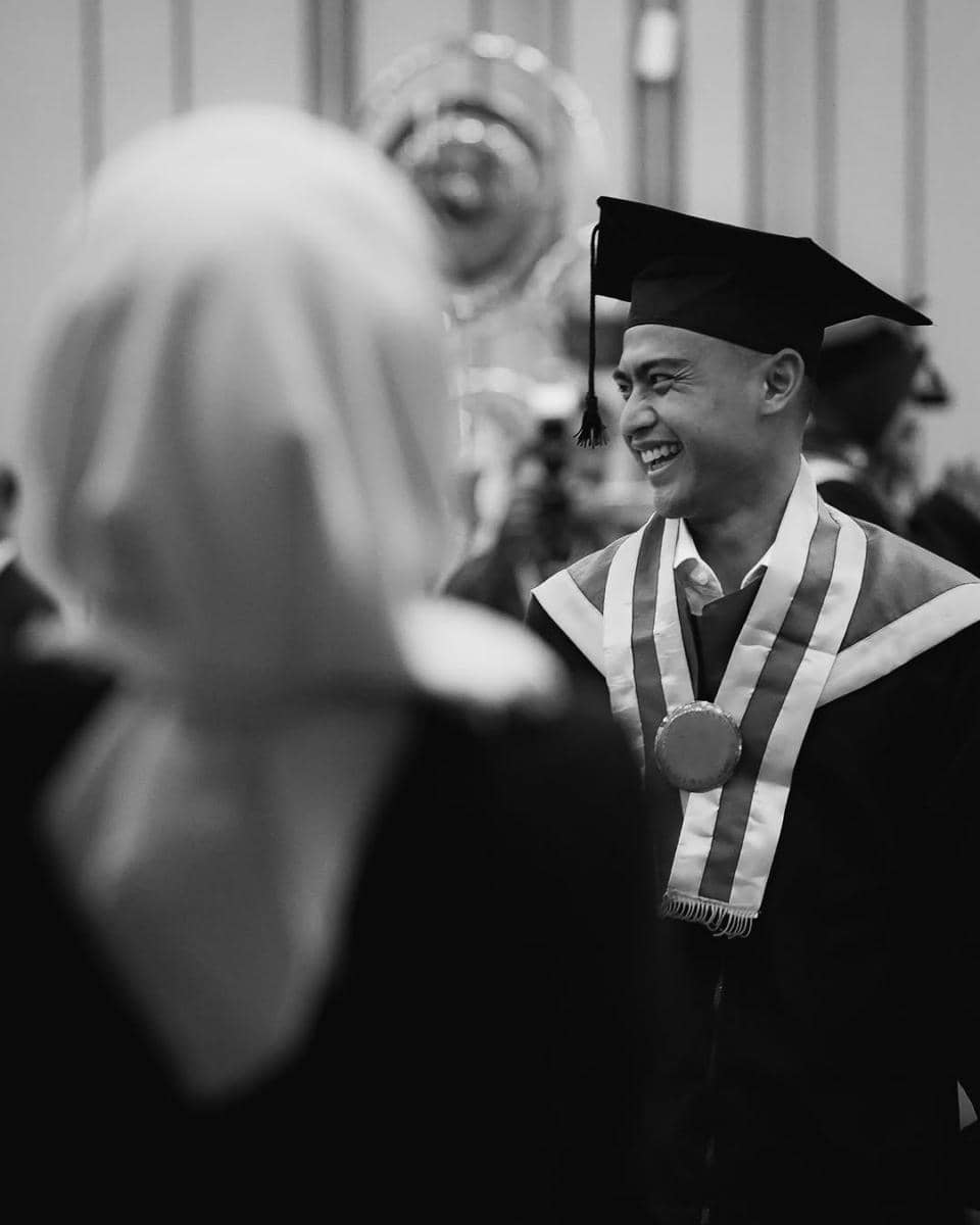 Pratama Arhan mengenakan toga dan medali wisuda tersenyum lebar di acara kelulusan, dengan seseorang berkerudung di depannya.