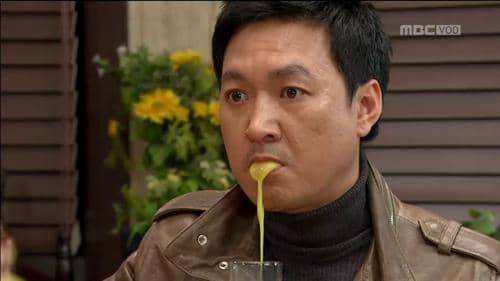 Seorang pria mengenakan jaket kulit cokelat dan sweter gelap sedang duduk di meja dengan cairan keluar dari mulutnya.