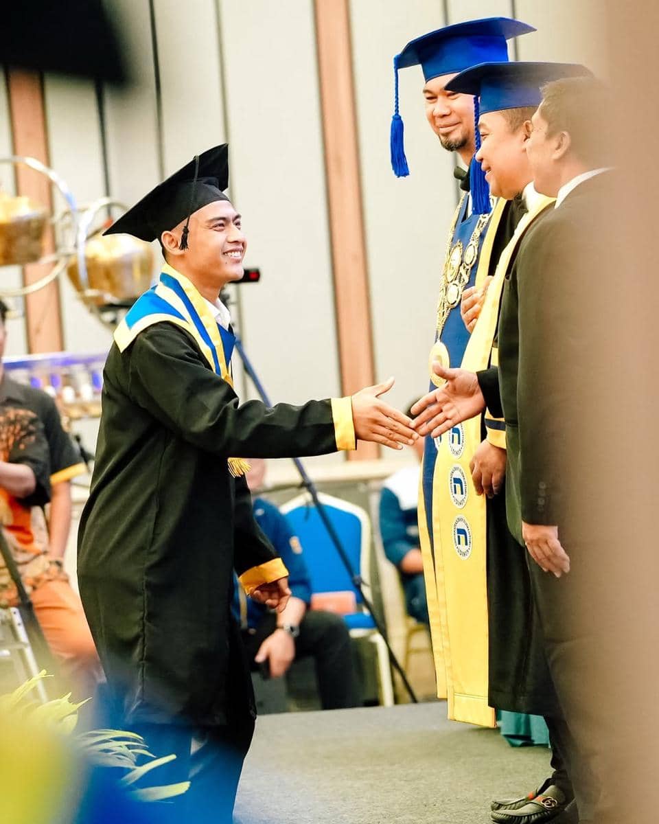 Pratama Arhan mengenakan toga hitam dan selempang biru emas berjabat tangan dengan dosen di atas panggung saat upacara kelulusan.