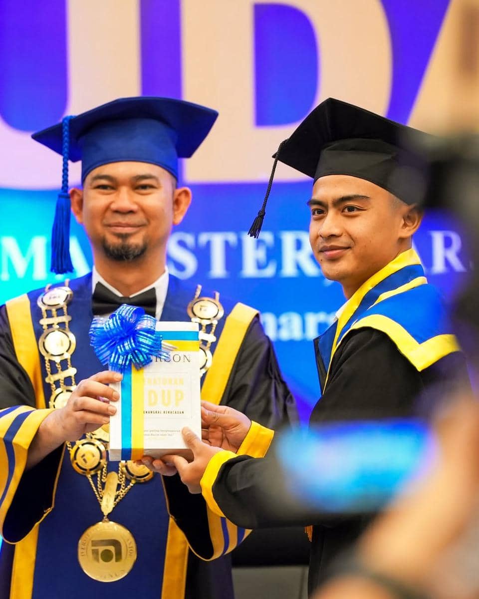 Seorang wisudawan mengenakan toga hitam dan biru menerima penghargaan dari dosen berpakaian toga biru dan kuning di acara wisuda.